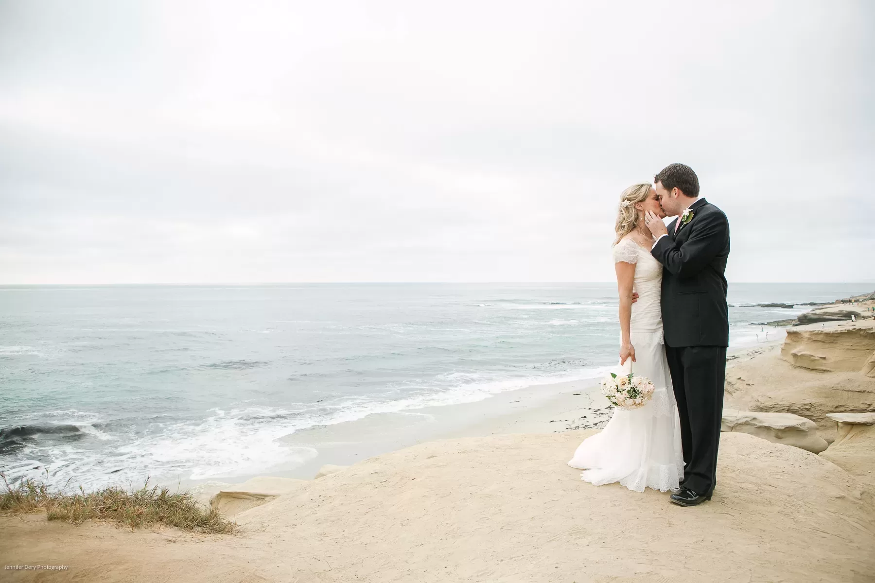 A bride and groom stand on a sandy cliff overlooking a calm ocean. The bride, in a white gown, holds a bouquet and gazes lovingly at the groom, who is dressed in a black suit. They share an intimate moment with the serene coastline in the background.