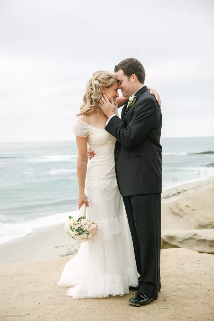 A bride and groom stand on a sandy beach with the ocean in the background. The bride, in a white wedding dress, holds a bouquet of flowers while the groom, in a black suit, gently touches her face. Both are smiling and sharing an intimate moment.