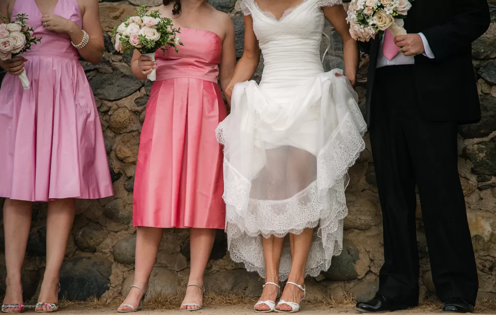 A bride in a white wedding dress stands with three attendants against a stone wall. Two bridesmaids are wearing pink dresses and holding bouquets, while the best man, dressed in a black suit with a pink vest, holds a bouquet as well. Faces are not shown.