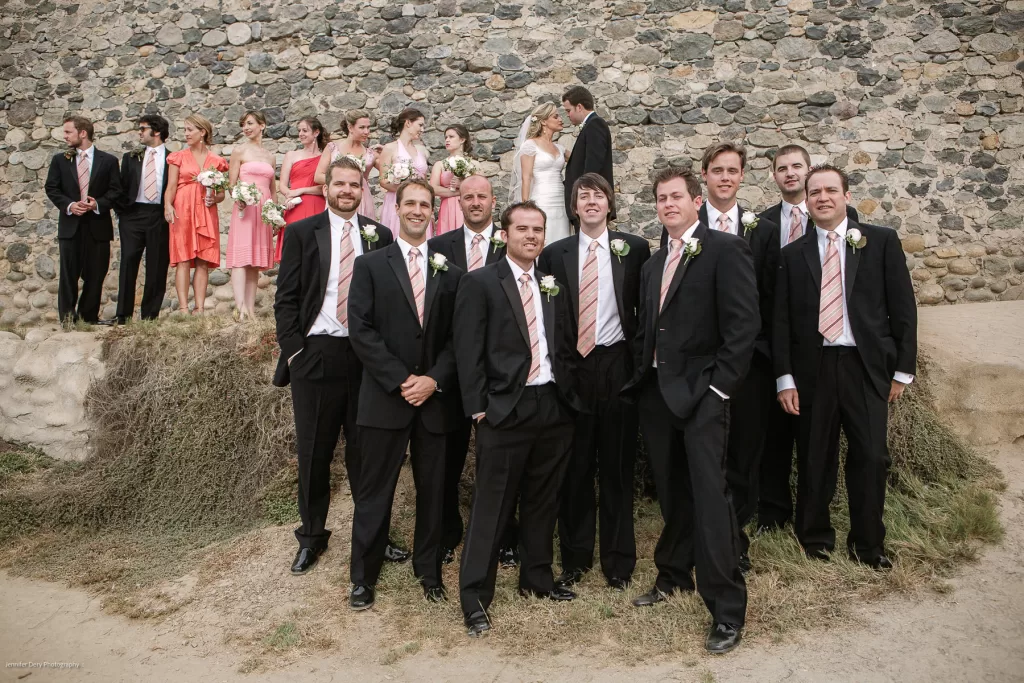 A wedding party poses for a photo. In the foreground, nine groomsmen wearing black suits, white shirts, and light pink ties stand on the ground. In the background, the bride, groom, bridesmaids, and a groomsman stand on a stone ledge.