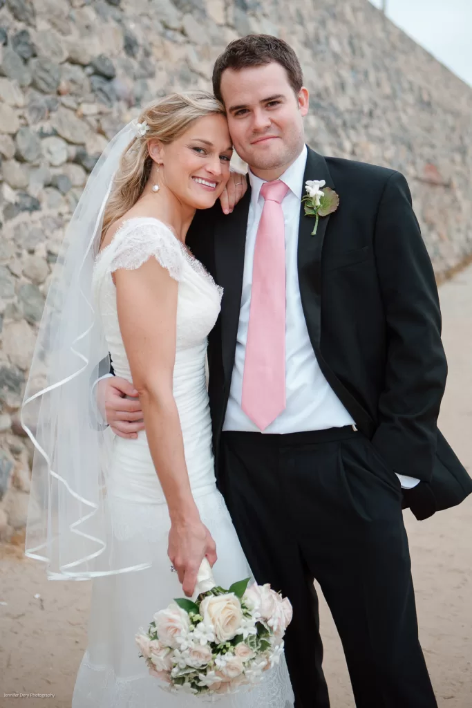 A bride in a white dress and veil stands next to a groom in a black suit with a pink tie. The bride holds a bouquet of flowers, and both are smiling. They pose in front of a stone wall on a sandy path. The groom has one hand in his pocket.