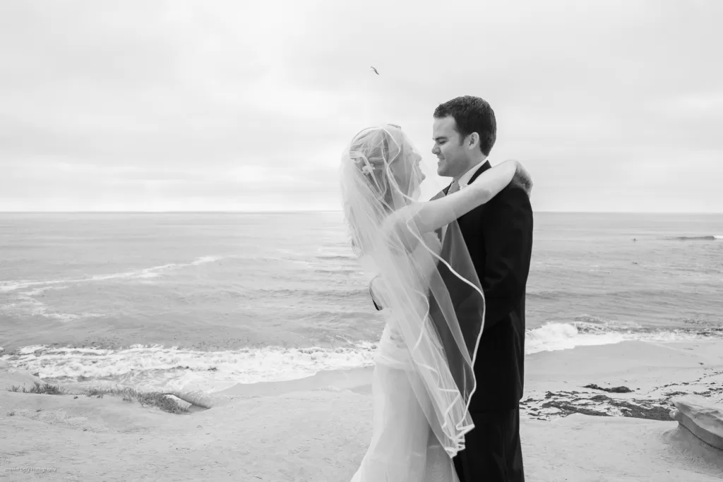 A black and white photo shows a bride and groom embracing on a cliffside overlooking the ocean. The bride wears a flowing dress with a long veil, and the groom is in a dark suit. Waves crash gently against the shore below, and the sky is overcast.