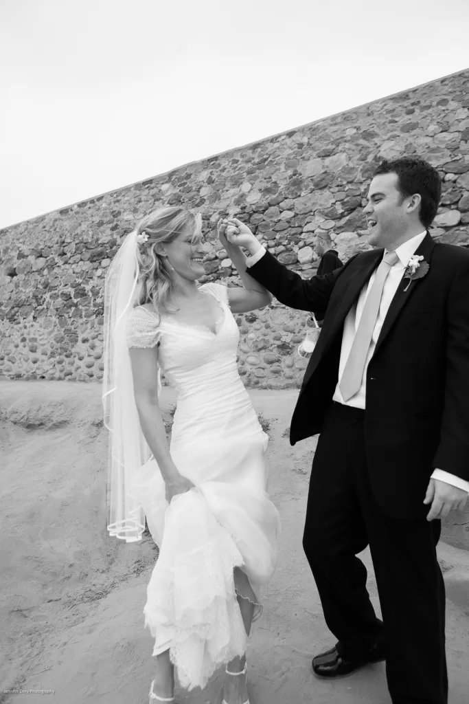 A black-and-white image of a bride and groom dancing on a sandy surface in front of a stone wall. The bride, wearing a flowing gown and veil, smiles as the groom, dressed in a suit, twirls her with his hand raised. Both appear joyous and carefree.