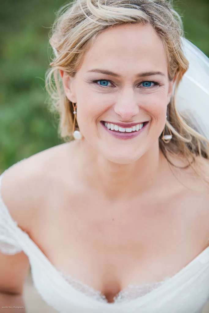 A smiling woman with blue eyes and blonde hair wearing a white bridal gown with lace details. She is also wearing drop earrings and a white veil. The background appears to be outdoors with greenery.