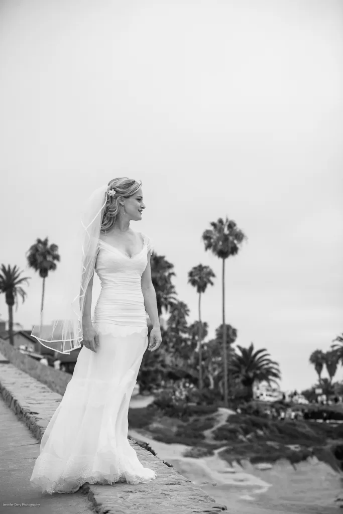 A bride in a flowing white gown and veil stands on a stone path overlooking a scenic, palm-tree-lined coastal view. Her gaze is directed towards the horizon, and the backdrop features a cloudy sky and lush vegetation. The setting exudes a serene, romantic atmosphere.