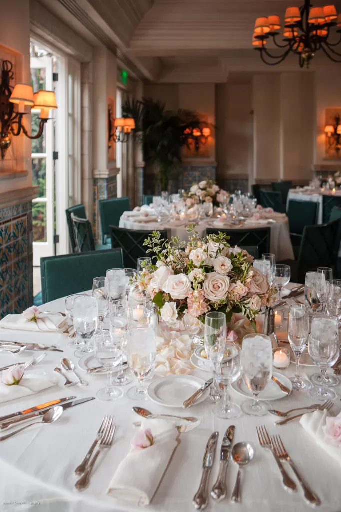 A beautifully arranged wedding reception table with white linens, elegant glassware, and cutlery. The centerpiece features a lush bouquet of white and pale pink roses with greenery. The room is warmly lit with chandeliers and wall sconces, creating an intimate ambiance.