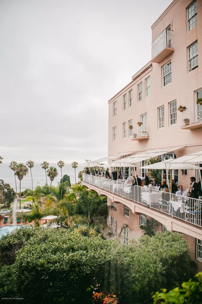 A large, elegant pink building with balconies overlooking a lush garden and a pool area. People are seen dining on a terrace with white tablecloths under a canopy. Tall palm trees in the garden with a cloudy sky and the ocean visible in the background.