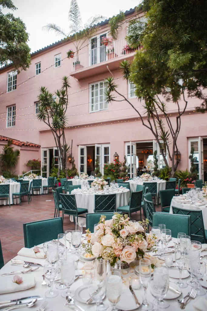 An elegant outdoor wedding reception setup with round tables covered in white tablecloths, green chairs, and floral centerpieces featuring white and pink roses. The venue is a pink building with balconies, tall windows, and string lights creating a romantic atmosphere.