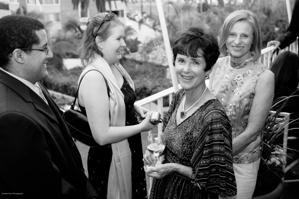 A group of four people socializing outdoors at a gathering. Three women and one man are smiling, dressed in semi-formal attire, and holding drinks. The background features greenery and a building. The atmosphere appears friendly and relaxed.