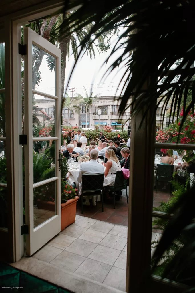 A view through open double doors looking out onto an outdoor wedding reception. Guests are seated at tables, engaged in conversation. The scene is surrounded by tropical plants and flowers, adding to the festive atmosphere beneath a partly cloudy sky.