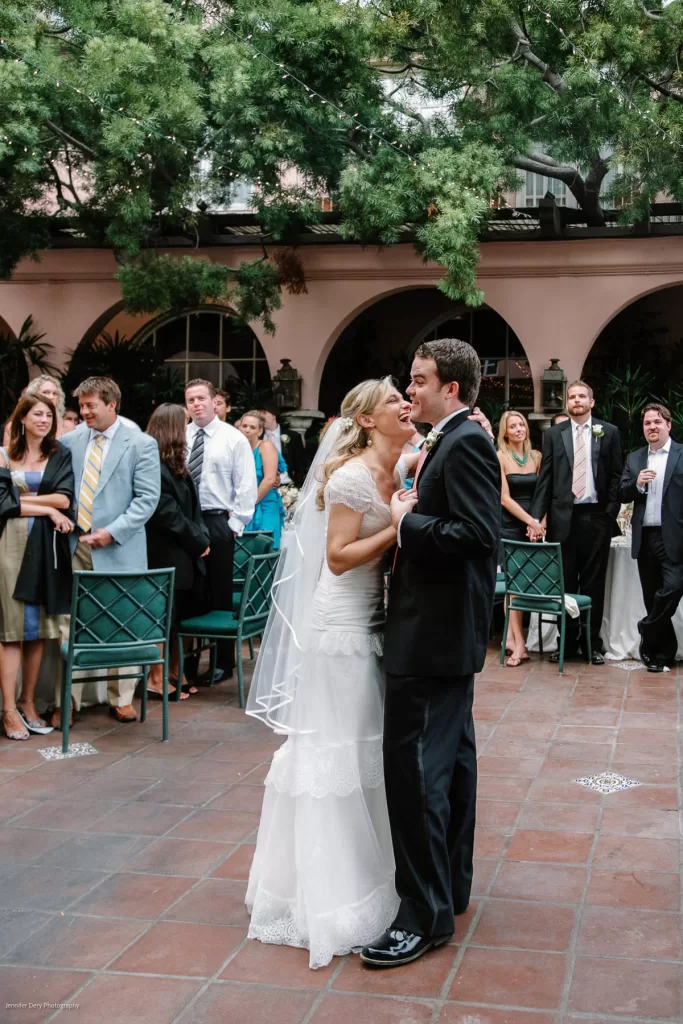 A bride and groom share their first dance at an outdoor wedding reception. The bride wears a white gown and veil, while the groom is in a black suit. Guests, dressed formally, watch and smile in the background. The venue is adorned with lush greenery and arched windows.