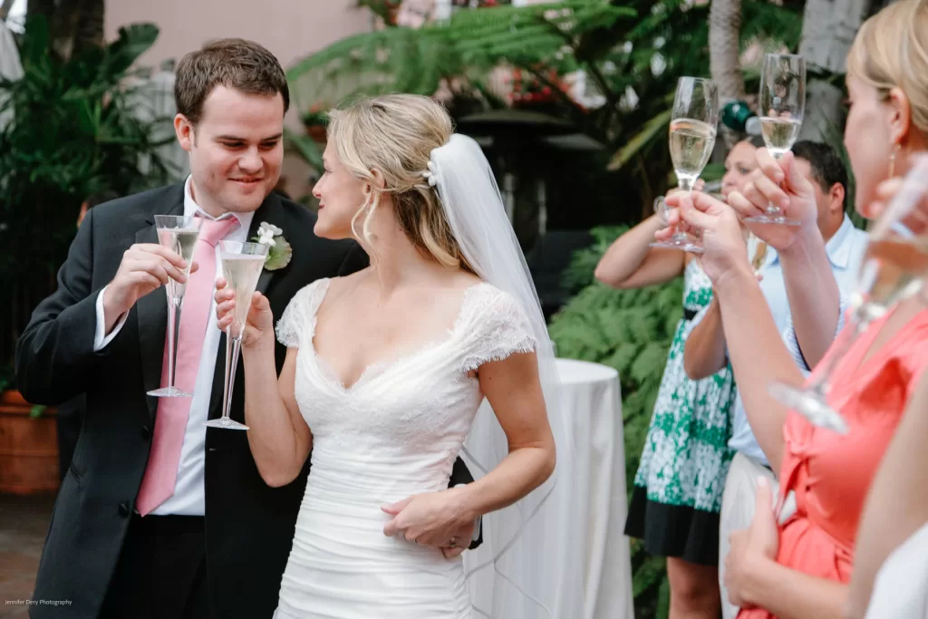 A bride in a white dress and veil clinks champagne glasses with the groom, who is wearing a suit with a pink tie. They are celebrating outdoors with guests who are also raising their glasses in a toast, surrounded by lush greenery.