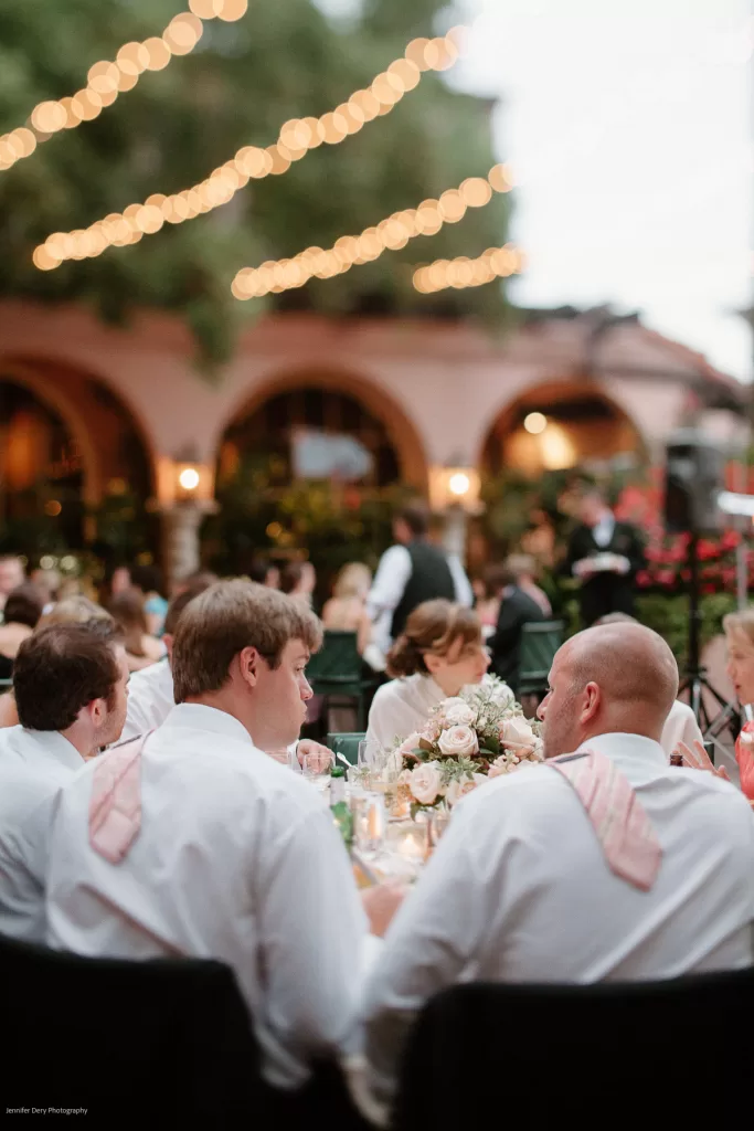 People dressed in formal attire sit at an elegantly decorated outdoor table under string lights. The background features blurred arched buildings with warm lighting, and a waiter in a black vest can be seen tending to a table. Comfortable and festive ambiance.