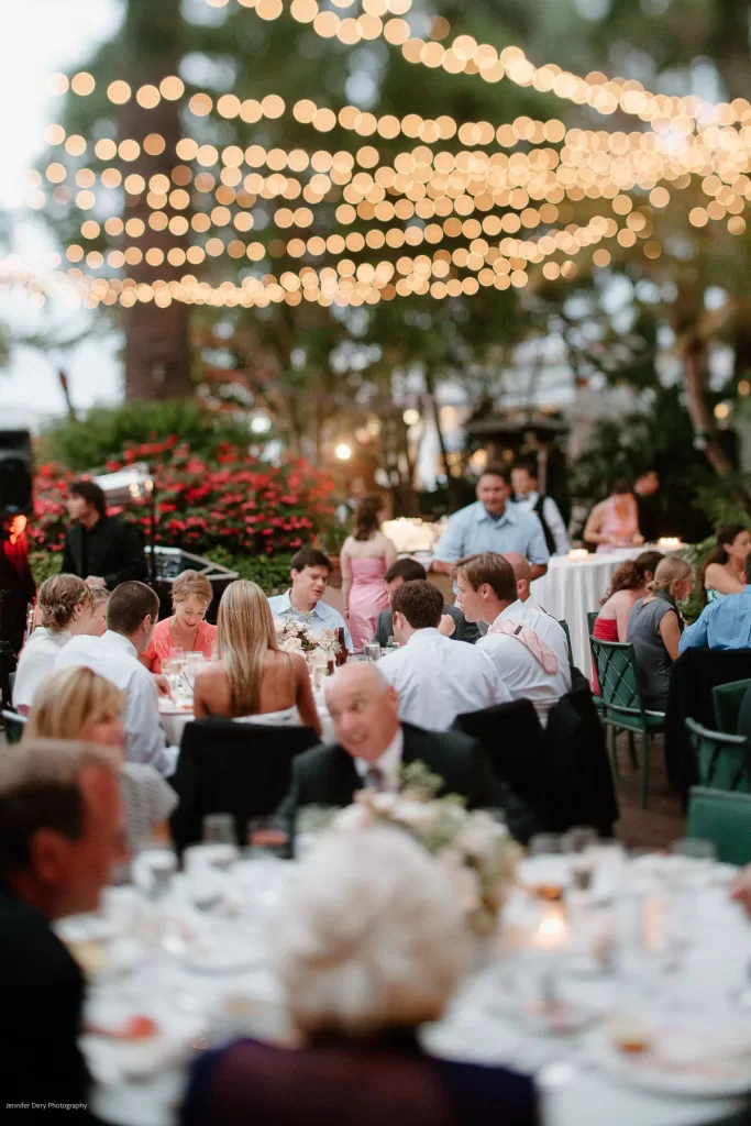 A lively outdoor wedding reception features guests seated at tables adorned with white tablecloths and elegant dinnerware. String lights are draped above, creating a warm, festive ambiance. The setting includes lush greenery and floral arrangements in the background.