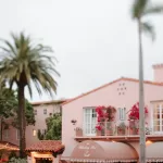 A quaint, pink two-story building with potted plants and vibrant red flowers. It has balconies and an awning reading "Whaling Bar." Palm trees are visible around the building, evoking a tropical, serene atmosphere under a cloudy sky.