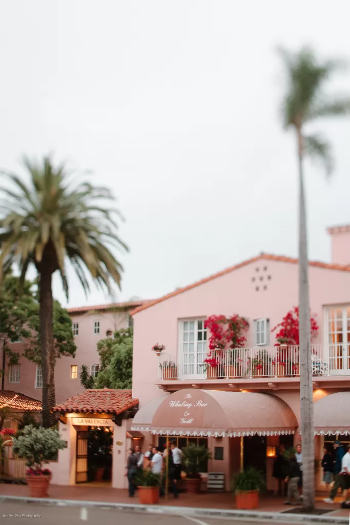 A quaint, pink two-story building with potted plants and vibrant red flowers. It has balconies and an awning reading "Whaling Bar." Palm trees are visible around the building, evoking a tropical, serene atmosphere under a cloudy sky.