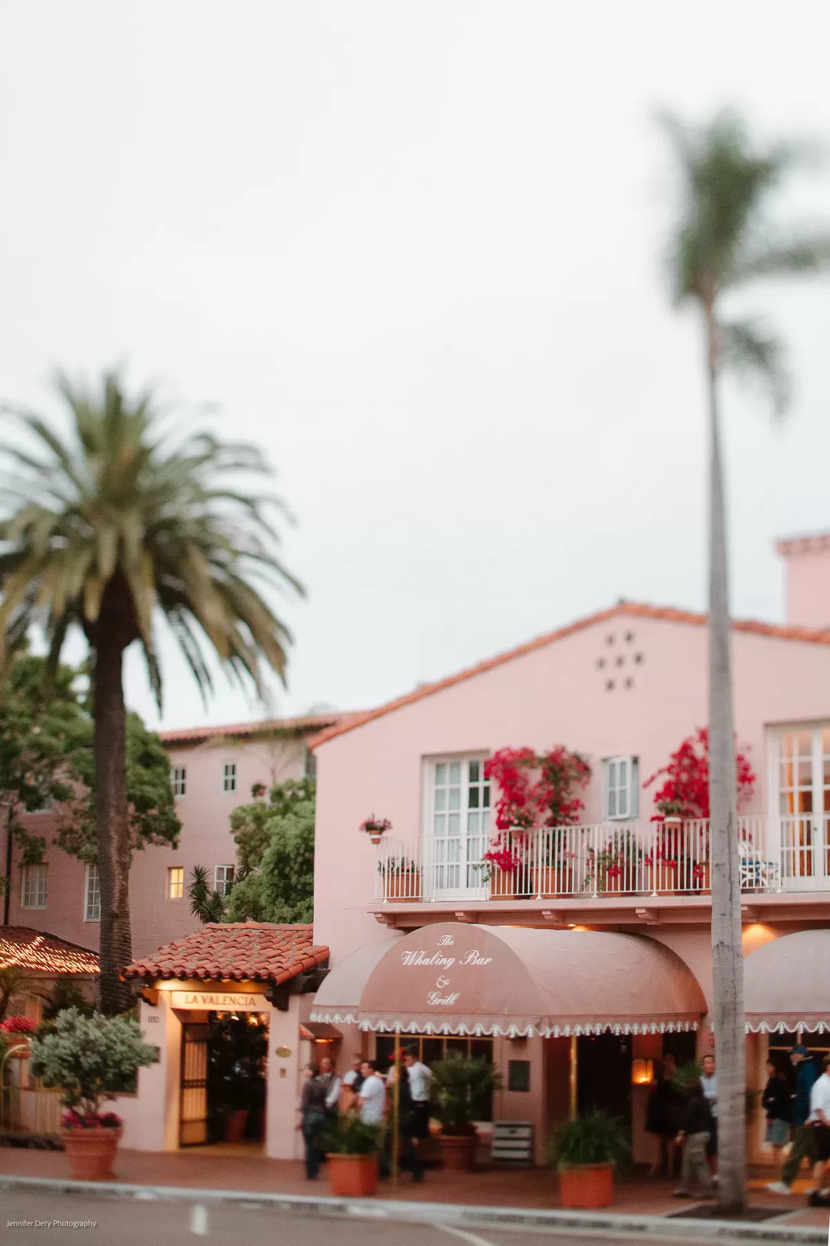 A quaint, pink two-story building with potted plants and vibrant red flowers. It has balconies and an awning reading "Whaling Bar." Palm trees are visible around the building, evoking a tropical, serene atmosphere under a cloudy sky.
