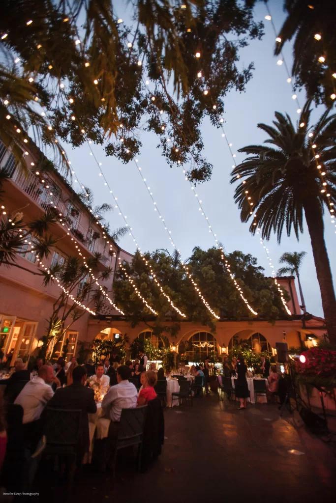 A lively outdoor evening event with people seated at tables, under a canopy of string lights. The venue is surrounded by palm trees and a building with balconies, creating a warm, festive ambiance.