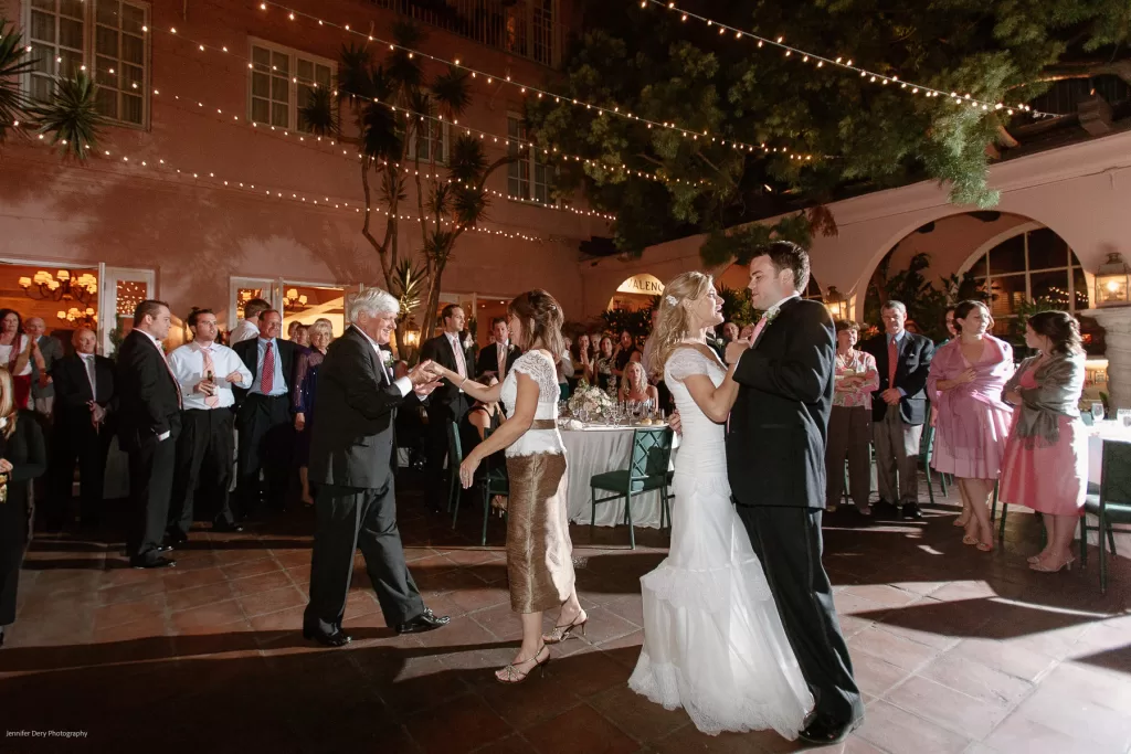 A wedding reception held outdoors in the evening; two couples dance on a tiled floor surrounded by guests. String lights drape overhead, creating a warm atmosphere. One couple includes a bride in a white dress and groom in a dark suit. The guests watch and smile.