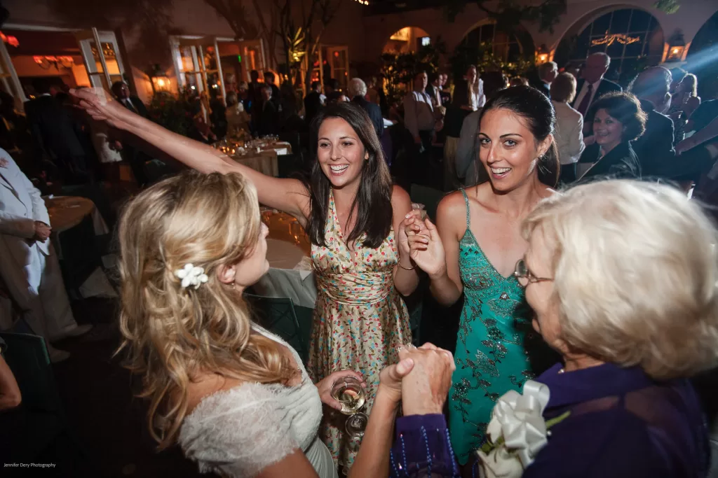 A group of four women, dressed in colorful attire, dance joyously at an indoor event. The woman in a light dress with flowers in her hair raises an arm while holding the hand of a woman in a green dress, as others around them laugh and engage in the celebration.