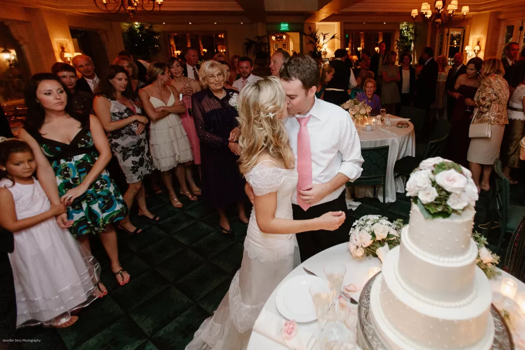 A bride and groom share a kiss in front of a three-tiered wedding cake in a warmly lit reception hall. Guests in formal attire surround them, some smiling and clapping. The room is decorated with flowers and elegant chandeliers.