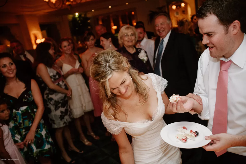 A bride and groom, both smiling, are celebrating at their wedding reception. The groom is holding a plate with a slice of cake, while the bride leans forward. Guests of various ages gather around them, appearing joyful and engaged in the moment.