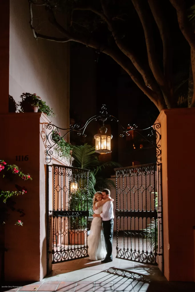 A newlywed couple embraces and kisses outside a wrought iron gate decorated with a hanging lantern. The scene is softly lit, highlighting the romantic atmosphere. The gate is flanked by stucco walls adorned with potted plants and the address "1116" is visible.