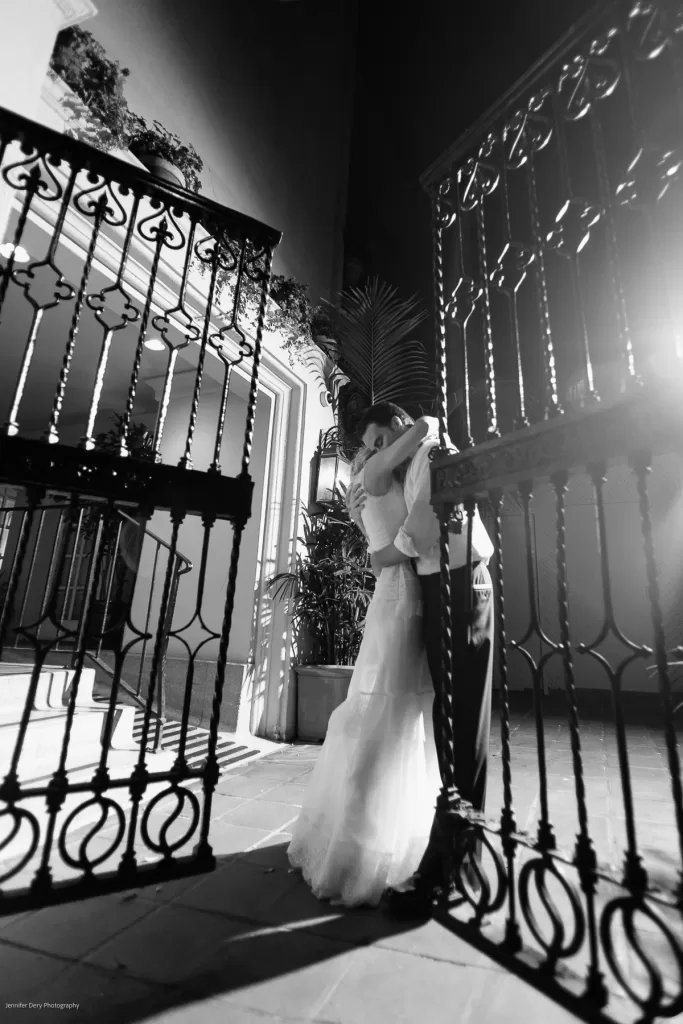 A black-and-white photo of a bride and groom embracing under an ornate wrought iron gate. The couple is backlit, creating a dramatic shadow effect. The bride wears a long gown, and the groom is in a suit. The setting appears elegant with architectural elements and plants.