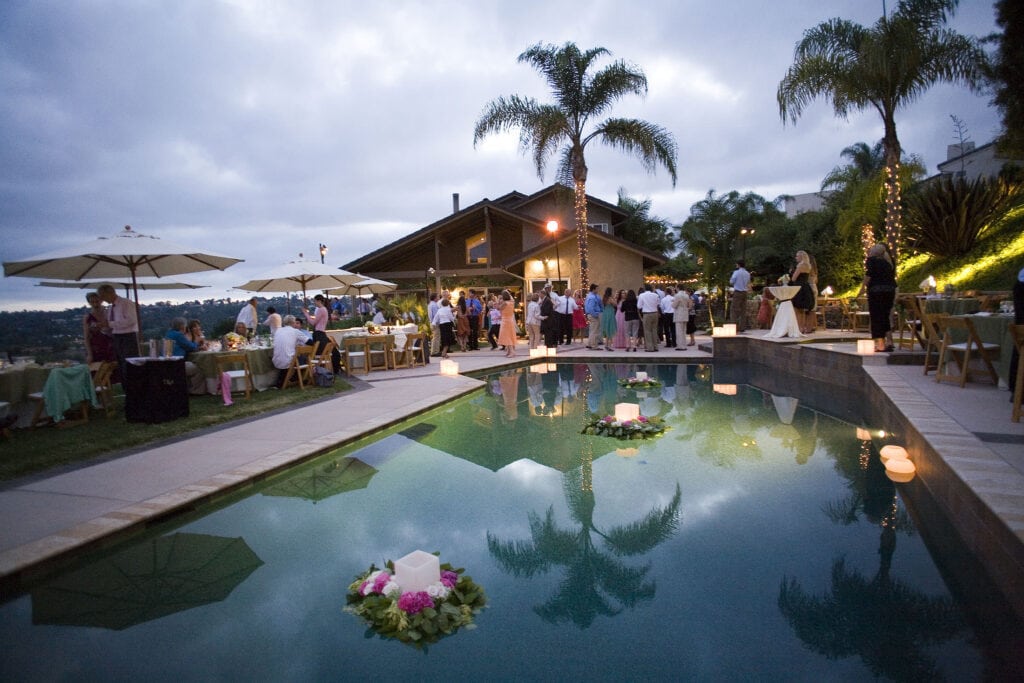 A poolside evening event with people mingling near tables under umbrellas. The pool has floating floral arrangements with candles. Palm trees and a house are in the background as clouds cover the sky. The scene is tranquil and festive, with light reflections on the pool water.