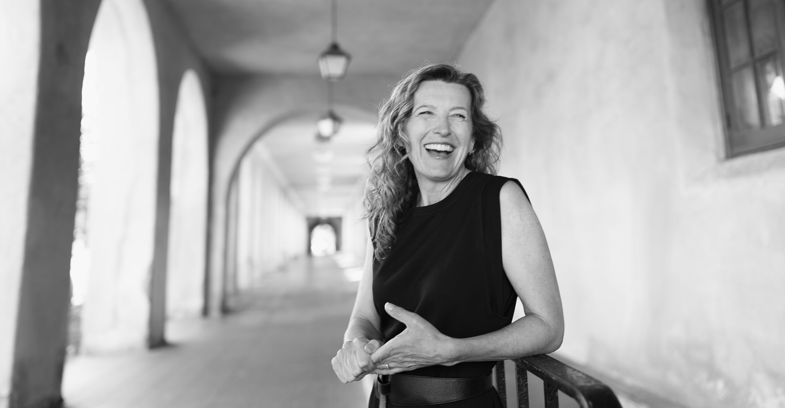 A woman with wavy hair, wearing a sleeveless black top, smiles and laughs as she stands in a long, arched corridor with hanging lights. She is near a railing, and sunlight filters through the arches. The image is in black and white.