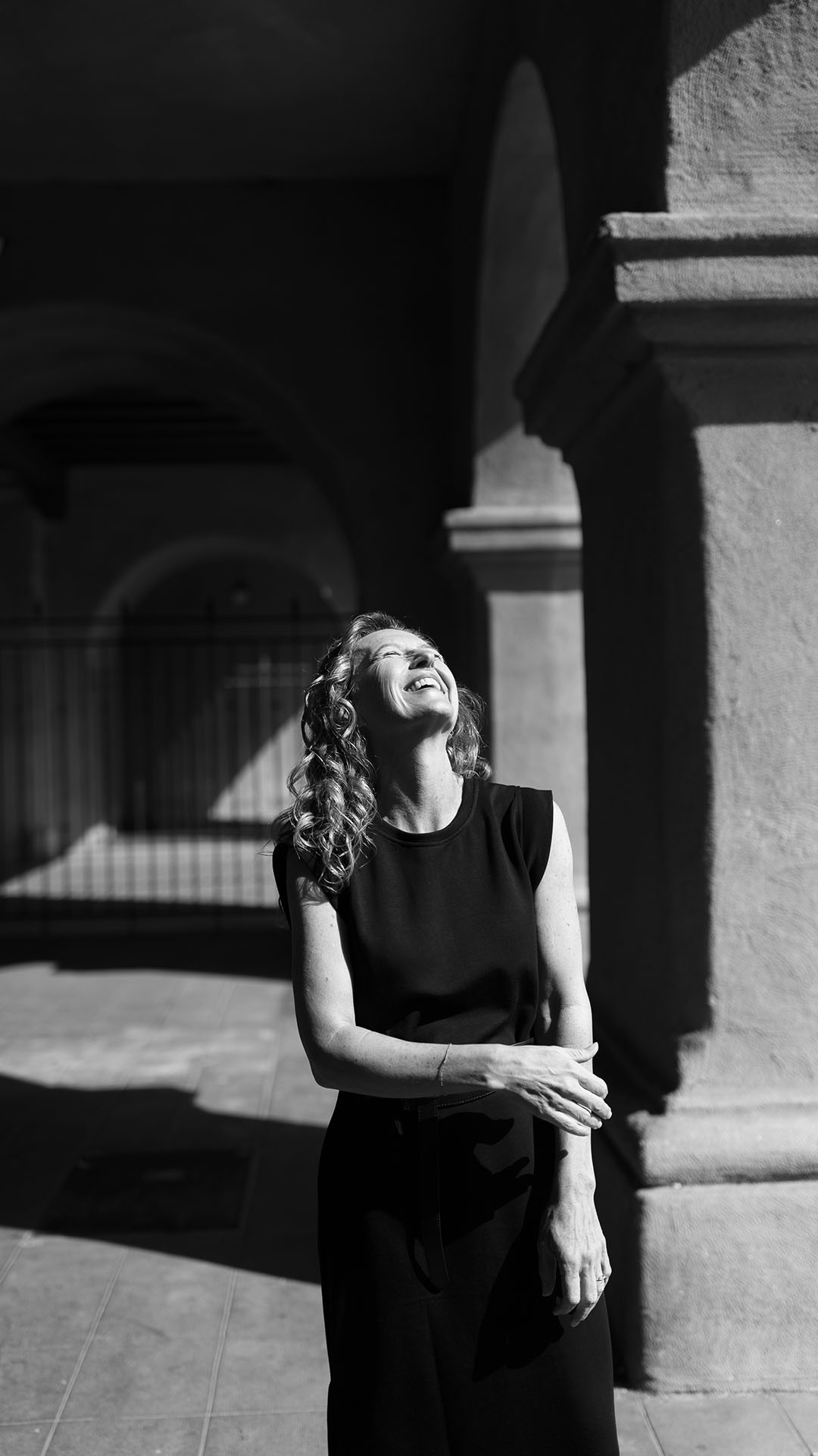 A woman with curly hair, wearing a sleeveless black dress, stands outside in front of an arched stone structure. Sunlight casts strong shadows as she looks upward and smiles, creating a striking contrast in the black and white photo.