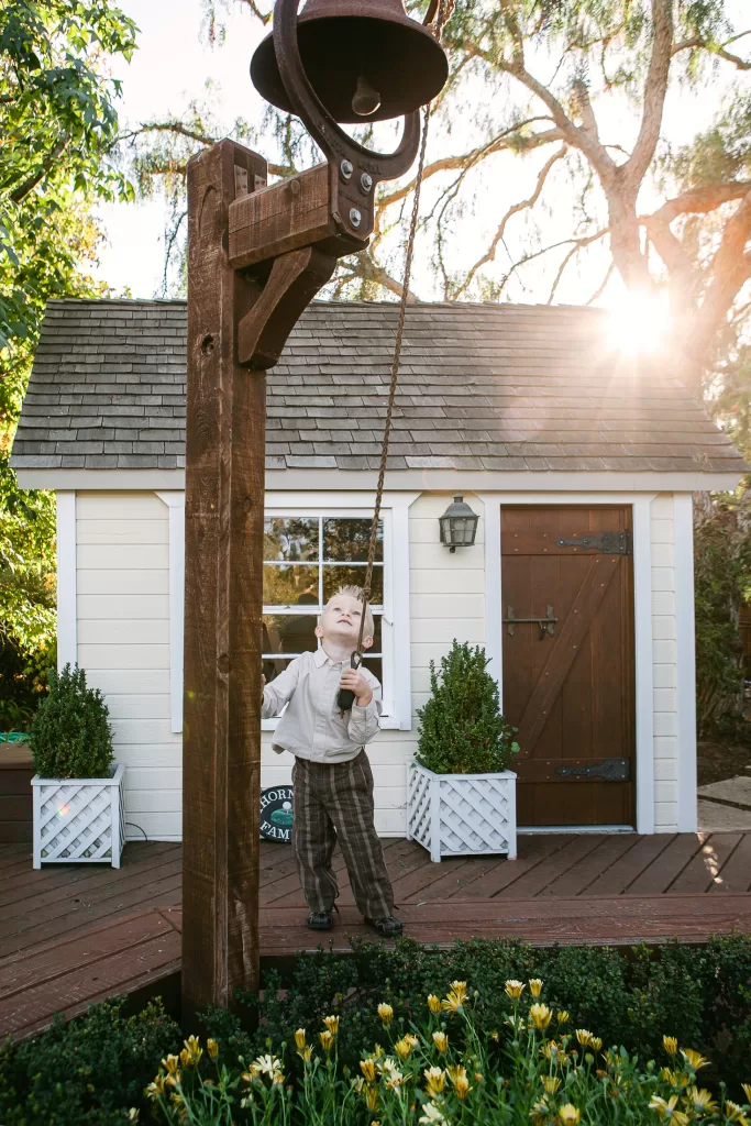 A young child wearing a light-colored shirt and plaid pants rings a large outdoor bell mounted on a wooden post. The child stands in front of a small white and brown playhouse, and the sun shines through trees in the background, illuminating the scene.