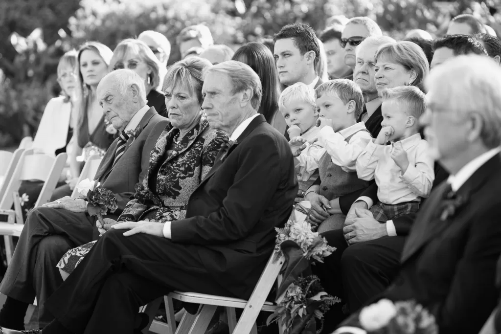 A black and white photo of an outdoor gathering. People are sitting attentively on chairs, dressed formally. In the foreground, a woman and a man appear focused. To their right, several children are also seated, with adults surrounding them. Trees are visible in the background.