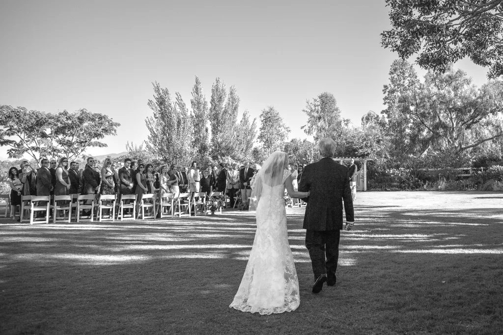 A bride in a white lace gown is walking down a grassy aisle with an older man, possibly her father, during an outdoor wedding. Guests are seated on white chairs, standing and watching as they pass by. Trees and greenery form a scenic backdrop under a clear sky.