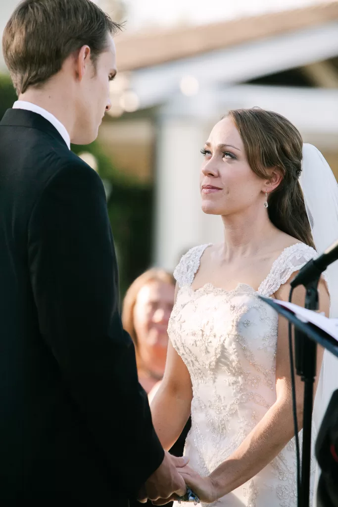 A bride and groom stand facing each other, holding hands during their wedding ceremony. The bride wears a lace wedding dress and a veil, looking up at the groom, who is dressed in a black suit and white shirt. Another person is visible in the background.