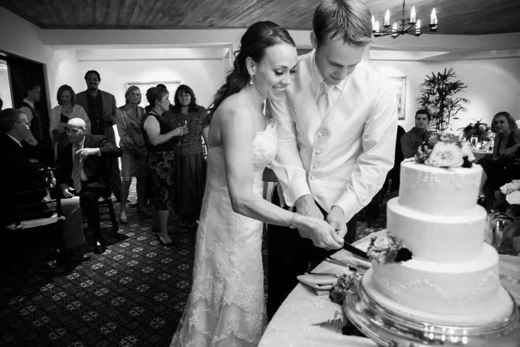 A bride and groom, dressed in wedding attire, are smiling as they cut a multi-tiered wedding cake together. Guests stand around them, watching and smiling. The scene is captured in black and white, adding a classic, timeless feel to the joyous moment.