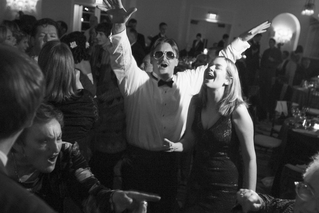A lively black-and-white party scene with a group of people dancing and singing. The man in the center wears sunglasses and a tuxedo, raising his arms, while a woman beside him in a dress is smiling. Other partygoers are visible in the background.