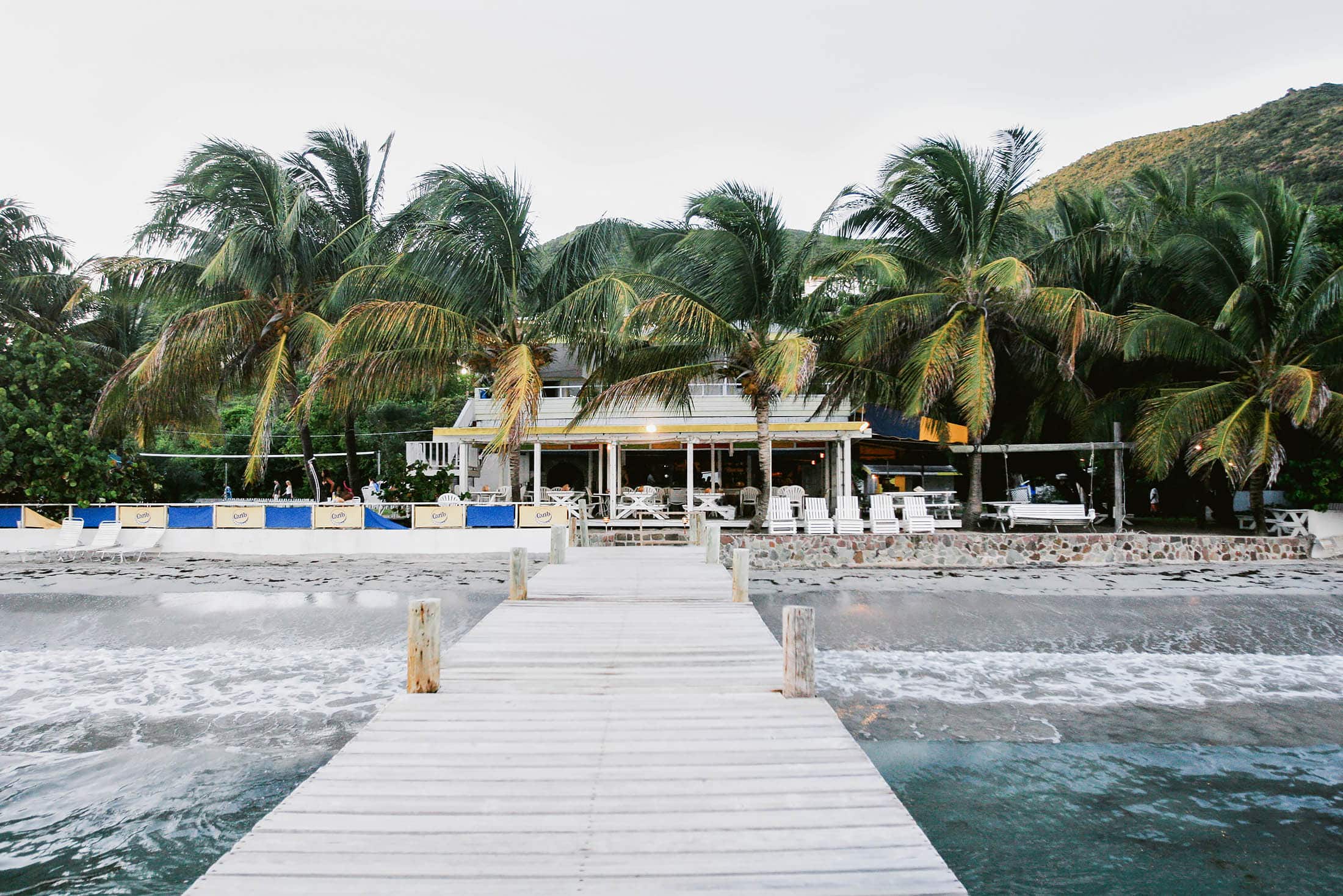 A wooden pier leads to a beachside restaurant surrounded by tall palm trees in Jamaica. There are tables and chairs on a deck, and gentle waves reach the sandy shore. A hill is visible behind the trees under a clear sky. Photographer specializes in Documentary Editorial Wedding Lifestyle 