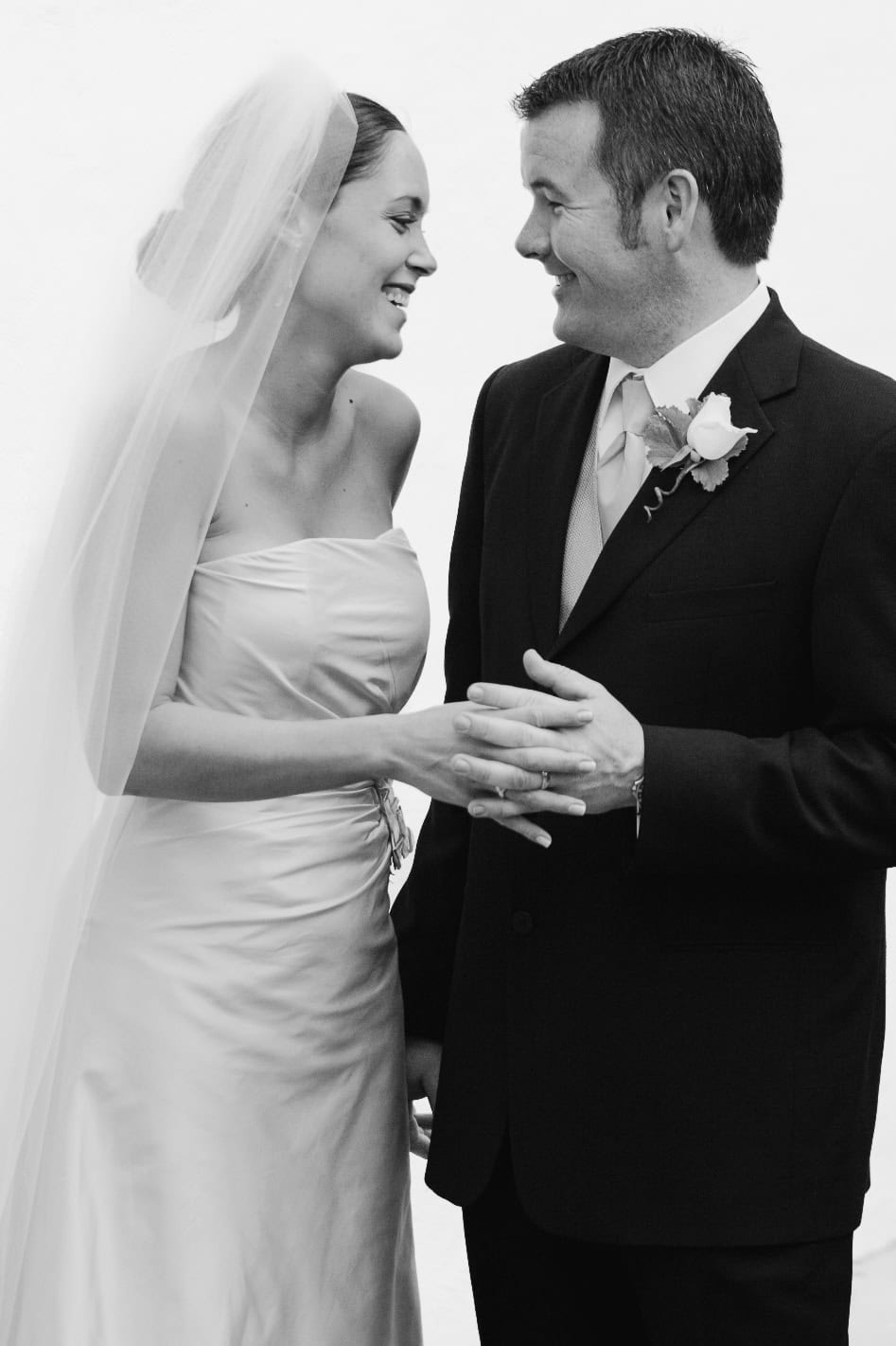 a bride and groom stand together, smiling at each other. The bride is wearing a strapless gown and veil, while the groom is in a suit with a boutonniere. They are holding hands and appear happy and joyful. Photographer specializes in Documentary Editorial Wedding Lifestyle 