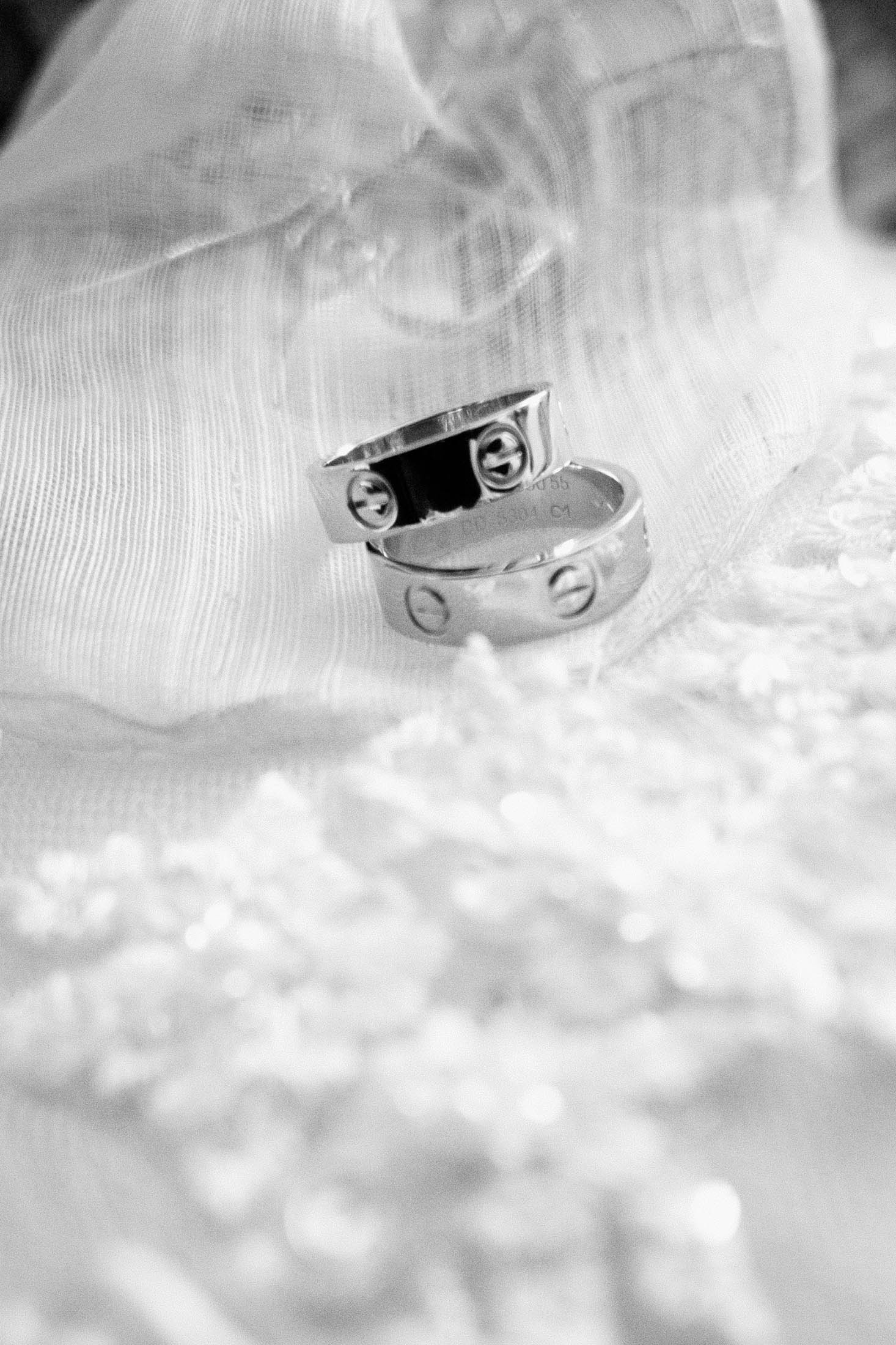 Two intertwined silver rings sit elegantly on a delicate piece of lace fabric, with a soft, sheer cloth draped around them. The rings feature subtle screw motifs, adding a modern touch to the classic scene.