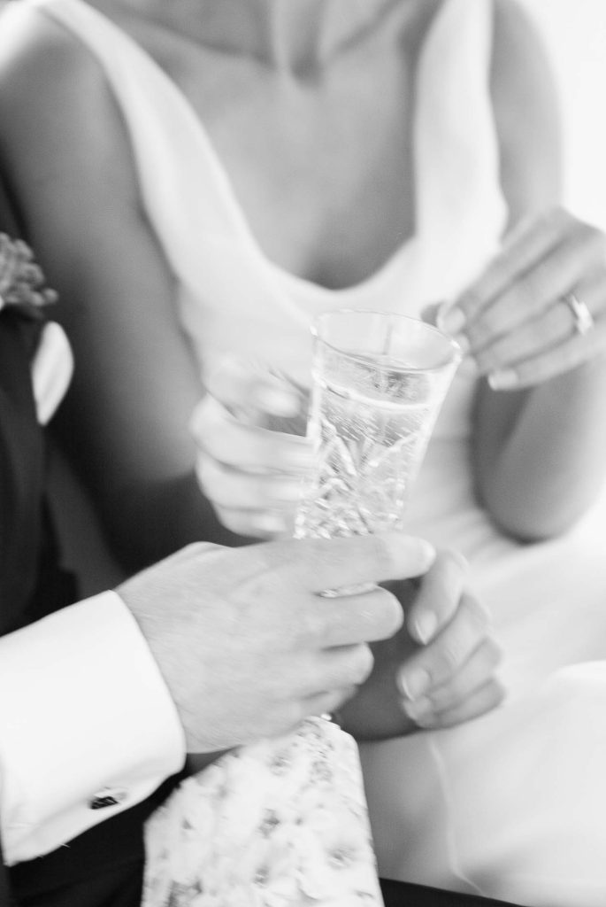A black and white photo shows a couple in formal attire holding a crystal champagne flute together, creating an intimate and celebratory atmosphere.