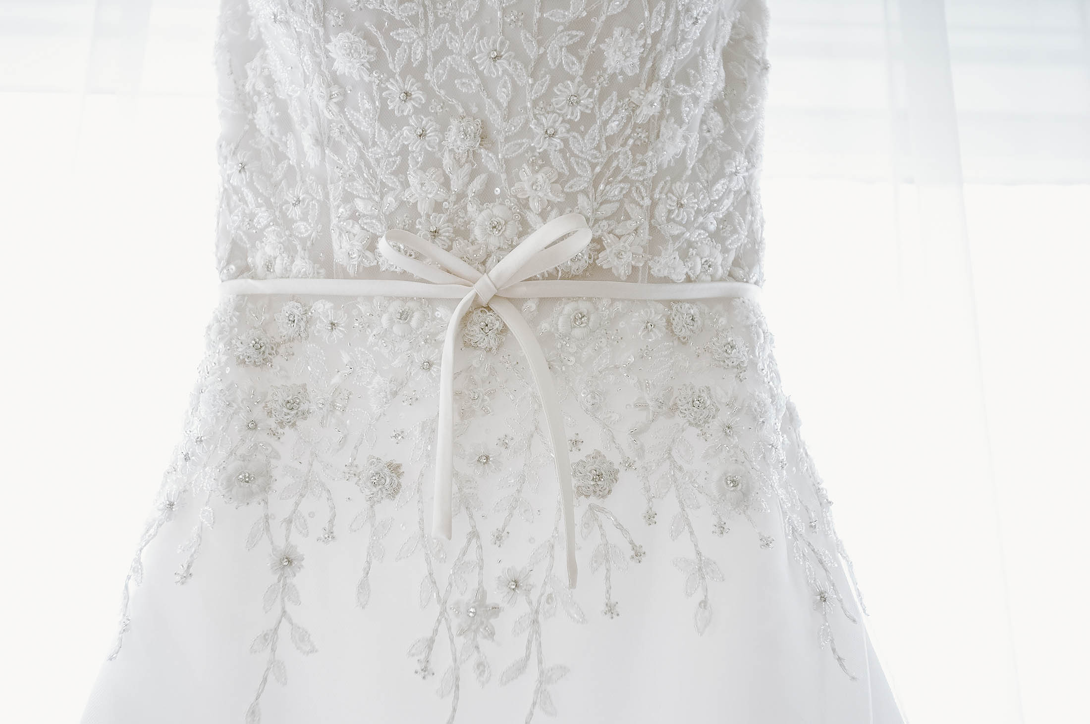 Close-up of an elegant white wedding dress adorned with intricate lace and floral embroidery. A delicate ribbon is tied into a bow at the waist, enhancing the intricate design. The background is softly lit, highlighting the dress's details.
