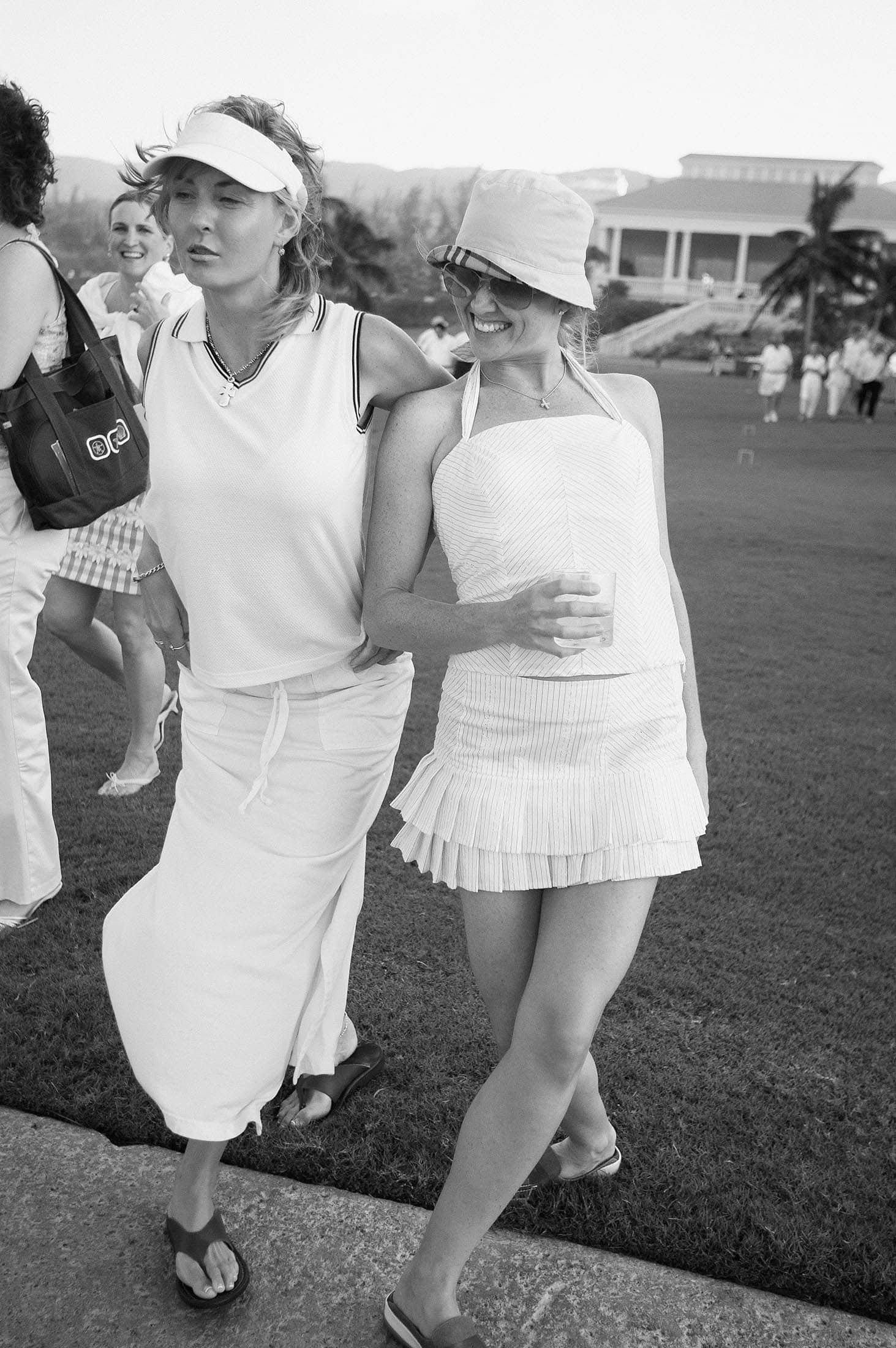 Two women in summer outfits walk arm in arm on a golf course in Jamaica. One wears a visor and long skirt, while the other wears a sunhat and short skirt. People and a large building are visible in the background. They're smiling and seem relaxed. Photographer specializes in Documentary Editorial Wedding Lifestyle 