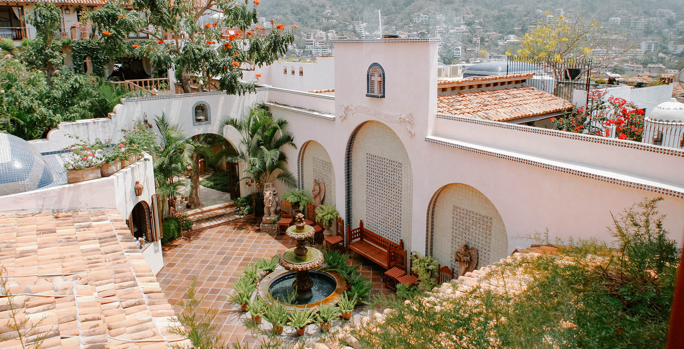 A picturesque courtyard with a central stone fountain surrounded by lush greenery, benches, and arched architectural features. The backdrop consists of traditional buildings and a scenic hillside view under a clear sky.