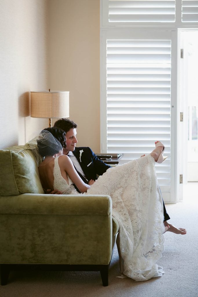 A couple dressed in wedding attire relaxes on a green couch in a softly lit room. The bride, wearing a white lace gown and birdcage veil, leans on the groom, who is in a black suit. Light filters in through white shutters.