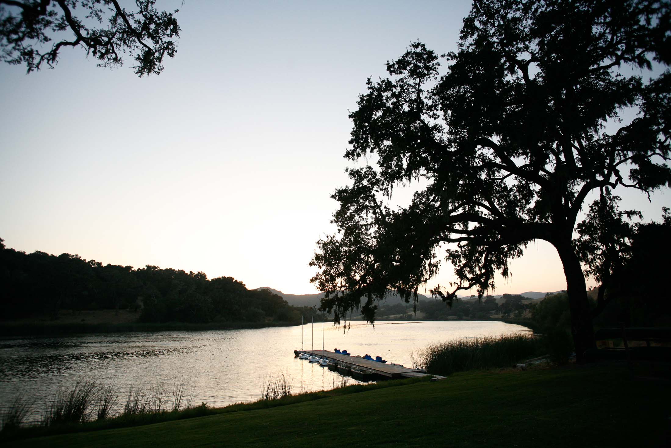 Silhouetted trees frame a serene lake at sunset, with a small dock and several sailboats anchored by the shore. The sky transitions from deep blue to soft orange, reflecting on the calm water.