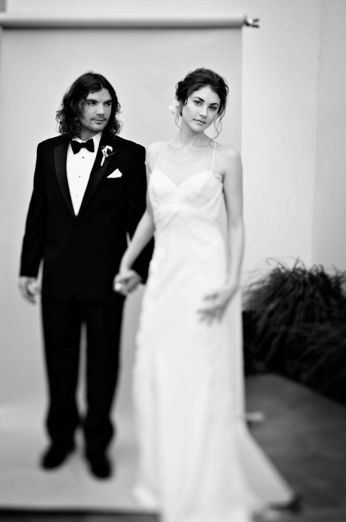A black and white photo of a couple dressed in wedding attire. The man is in a tuxedo, and the woman is in a long, elegant gown. They are holding hands and looking in different directions, standing against a plain backdrop.