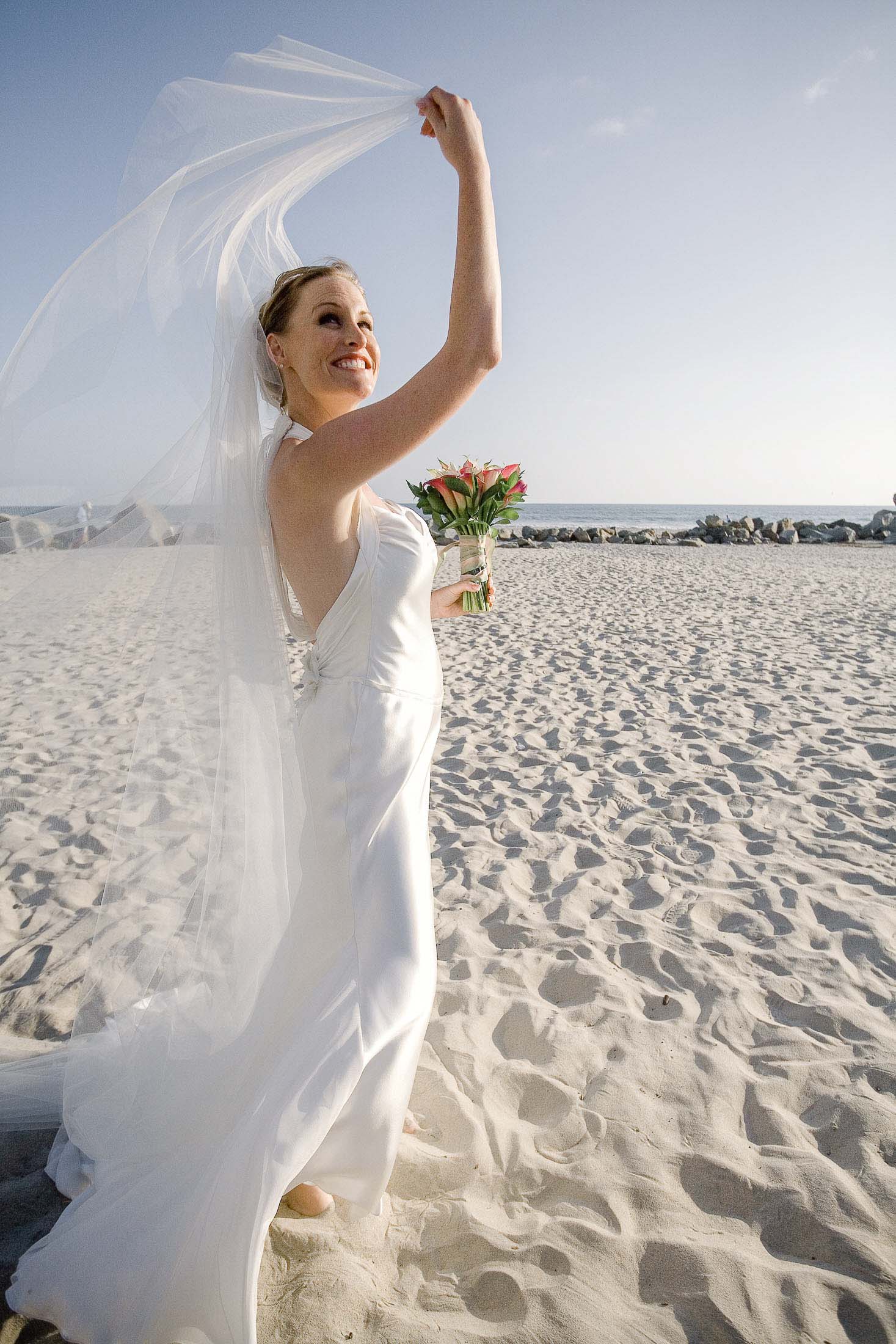 A bride in a flowing white gown and veil smiles and holds a bouquet of flowers on a sandy beach under a clear blue sky. Photographer specializes in Documentary Editorial Wedding Lifestyle 