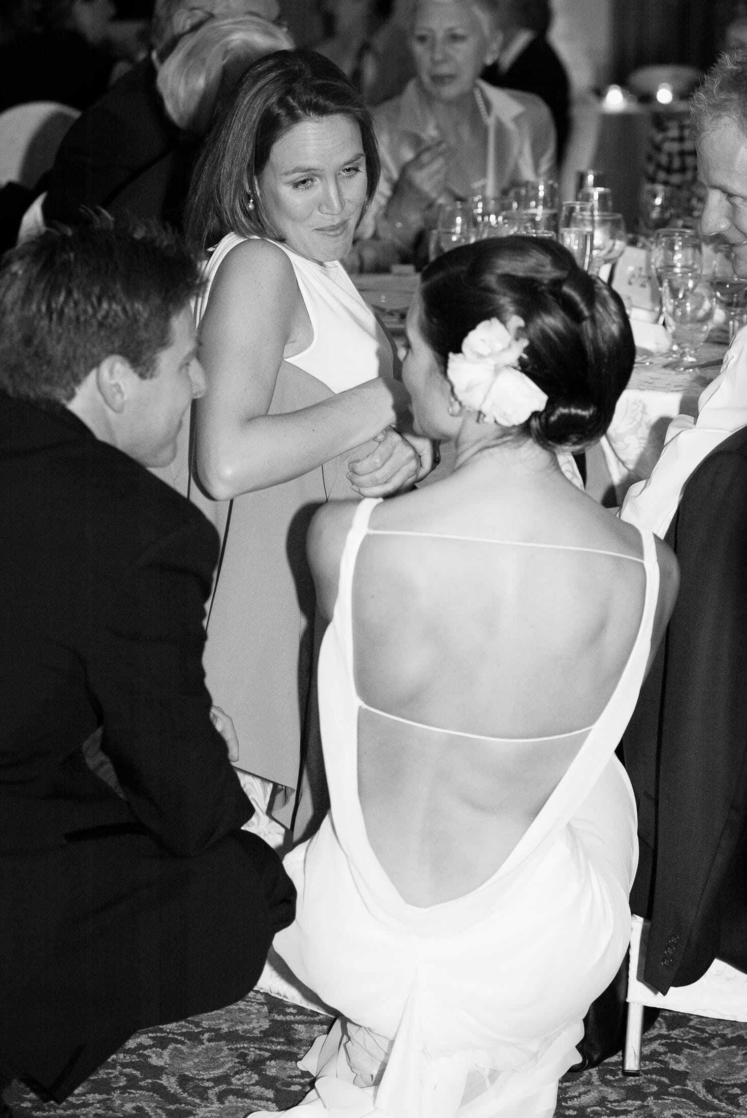 A black-and-white photo of people at a formal event. A woman in a backless dress is seated, facing a woman in a sleeveless dress. Two men in suits are nearby. They're engaged in conversation at a table with glasses and tableware. Photographer specializes in Documentary Editorial Wedding Lifestyle 