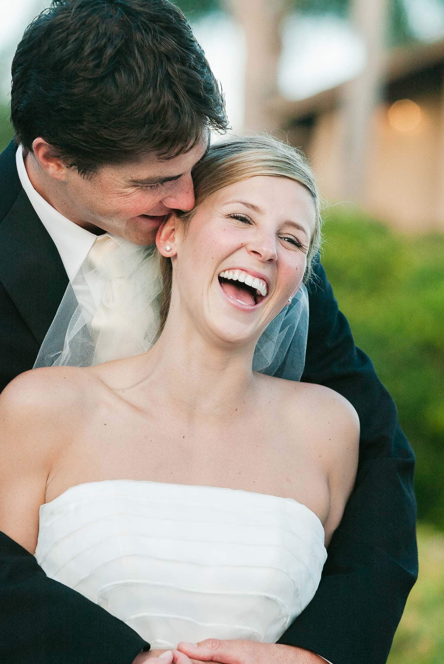 A groom in a black suit embraces a laughing bride in a white strapless wedding dress. They are outdoors with blurred greenery and a building in the background. The bride is smiling widely, displaying joy and happiness. Photographer specializes in Documentary Editorial Wedding Lifestyle 